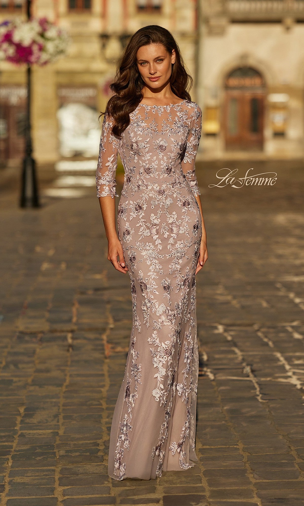 A woman wearing the La Femme 31464 long formal dress in mauve with three-quarter sleeves and light-colored floral lace embroidery stands on a cobblestone street.